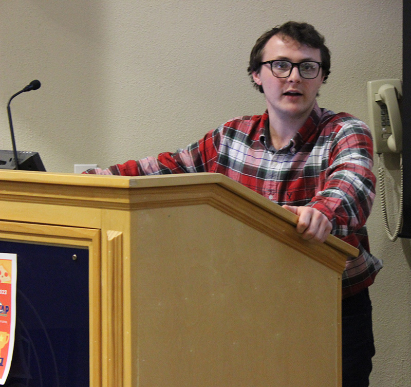 Male student with glasses and checked shirt making remarks while standing at a podium.