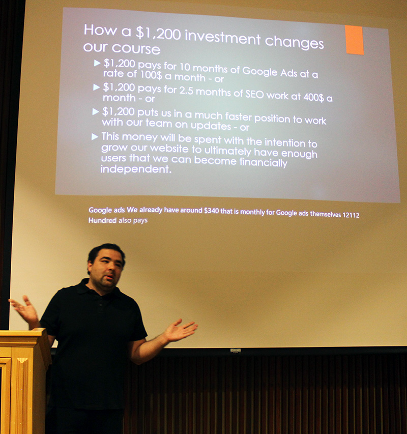 Male student with short dark hair and dark shirt standing in front of projection screen with a presentation slide.