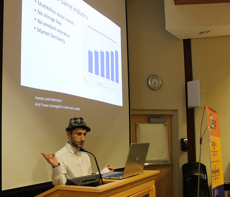 Student in White Shirt and Hat at podium with presentation slide on screen in background.