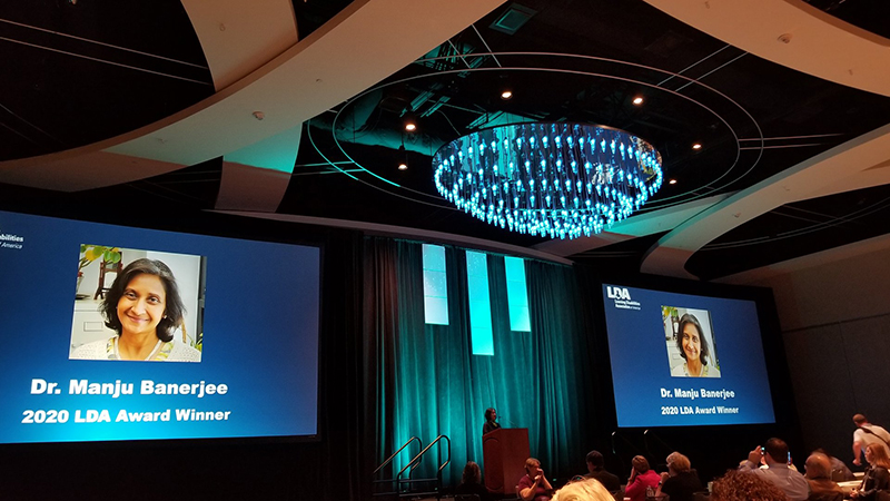 A long distance shot of Manju Banerjee speaking the podium in a ballroom at the LDA Conference in Orlando after accepting her award. Two video screens on each side her show her picture with text that reads Dr. Manju Banerjee 2020 LDA Award Winner.
