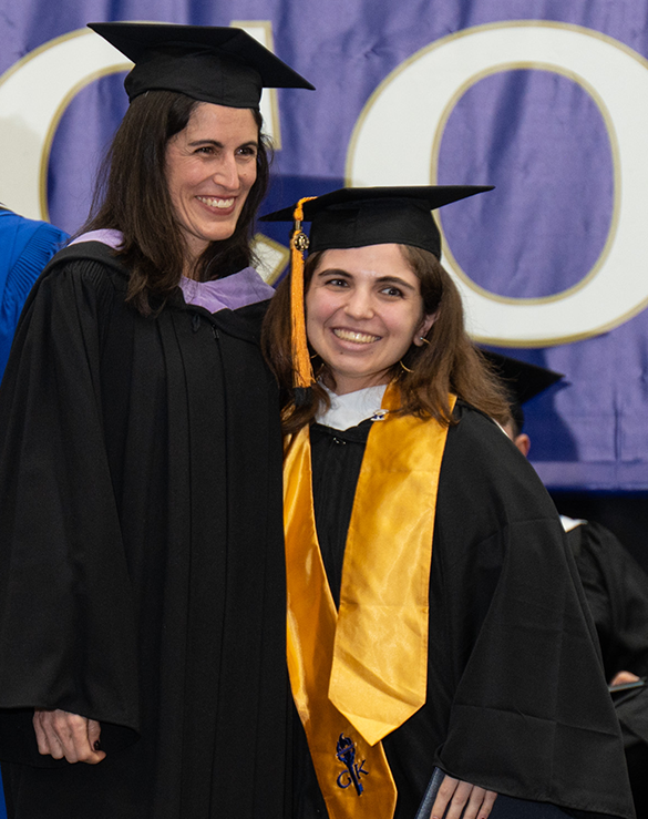 Two women in graduation regalia smile while posing for an on-stage photo during graduation.