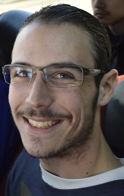 A close up of Micah looking at the camera and smiling. He has very short brown hair and a wispy mustache. He is wearing glasses and blue shirt on top of a white t-shirt.