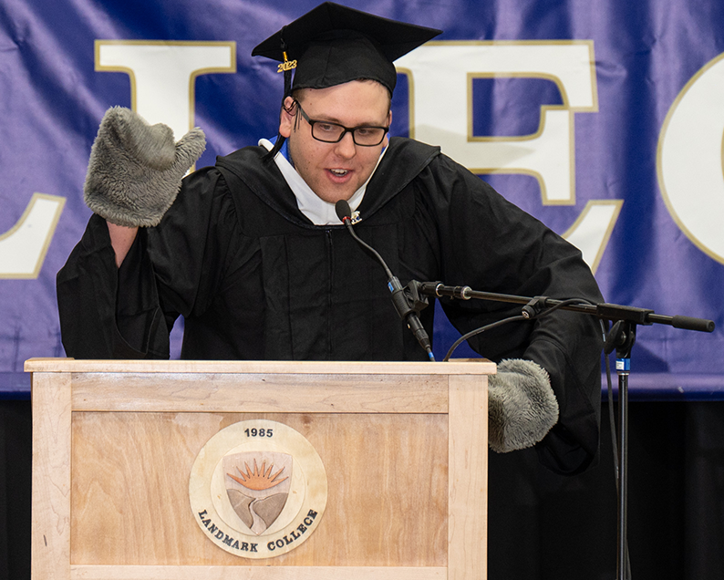 Male student with glasses and black graduation cap and gown stands a podium waving at audience with a gray flipper on his hand.
