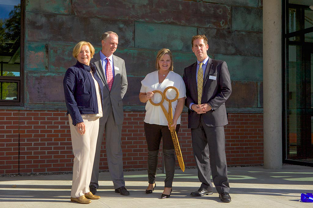 Four people standing in front of a building, with the woman in the middle holding a giant gold pair of scissors.