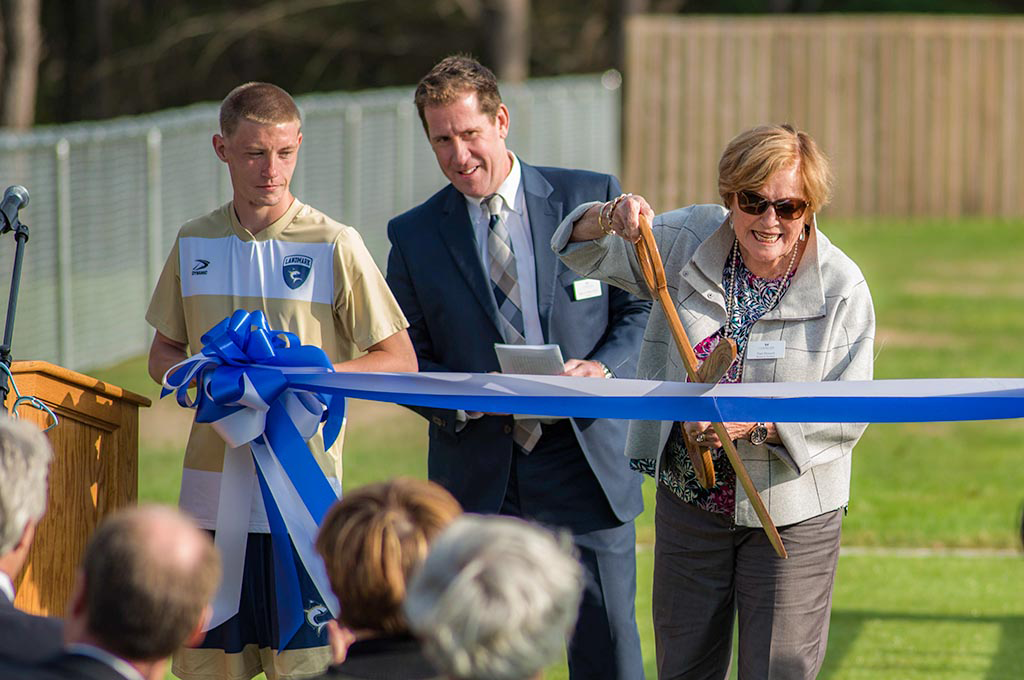 Peter Eden and a student watching Nan cut a large blue and white ribbon with giant gold scissors.