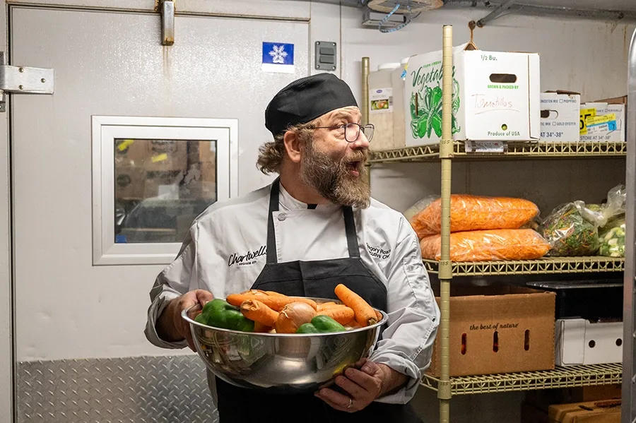 A chef makes a face of exaggerated surprise towards something off-camera, while holding a bowl of vegetables in an industrial kitchen.