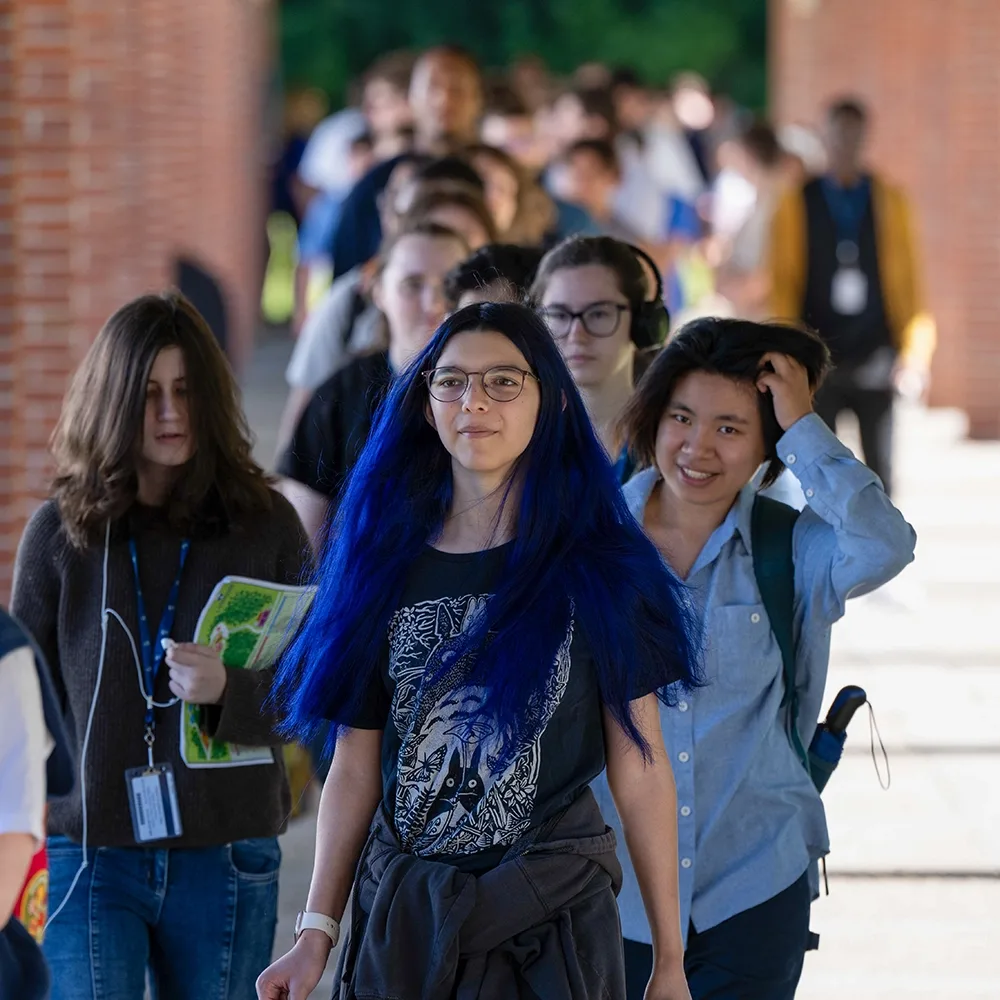 A group of students walk in a line down a colonnade.
