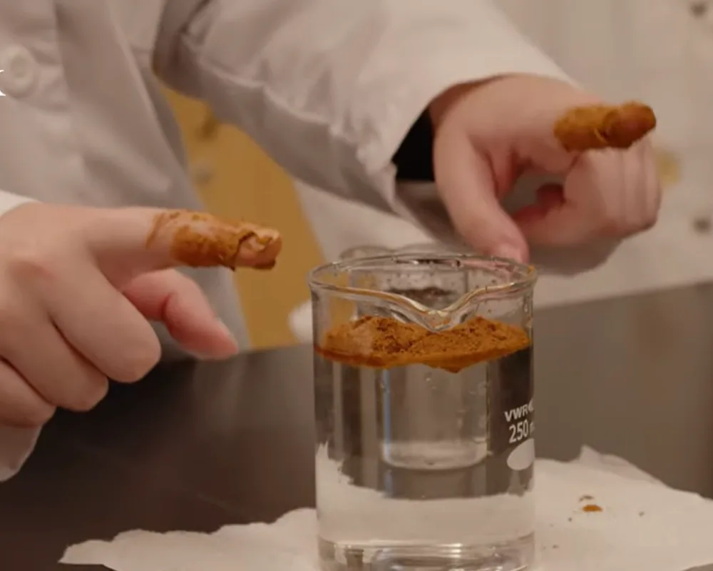 Close-up on two hands with index fingers covered in cinnamon and a large beaker of water on a table