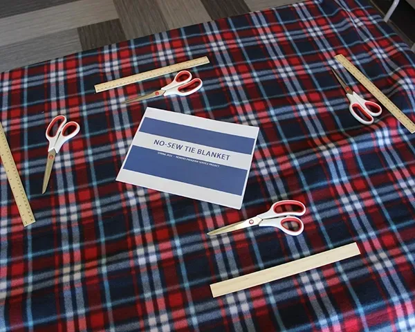 Plaid patterned fabric on a table with four rulers and pairs of scissors laid out along with a printed set of instructions for making a no sew tie blanket