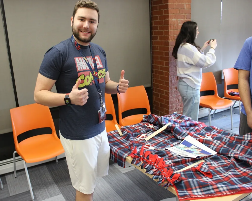 Male student in shorts and t-shirt giving thumbs up signal with hands as he stands next to the no sew blanket he is assembling.