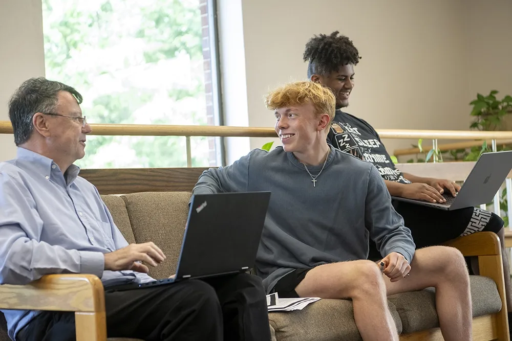 A professor and two male students sitting together on a couch and laughing about something.