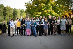 Twenty-five people stand outside to pose for a photo on a sunny day with trees and a brick building in the distance.