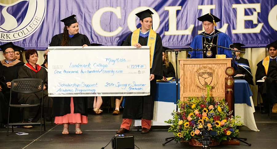 People in caps and gowns sit and stand on a stage. Two people hold up a May 14, 2016, check to Landmark College for $1279 from 2016 Spring + Summer Graduates for Scholarship Support and Academic Programming. The College president, in blue, speaks at a podium.