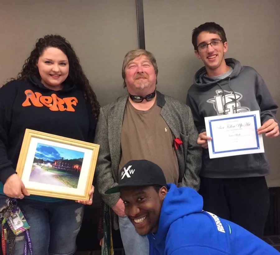 Four people pose for a photo. One holds a framed photo of a residence hall, and another holds a certificate.