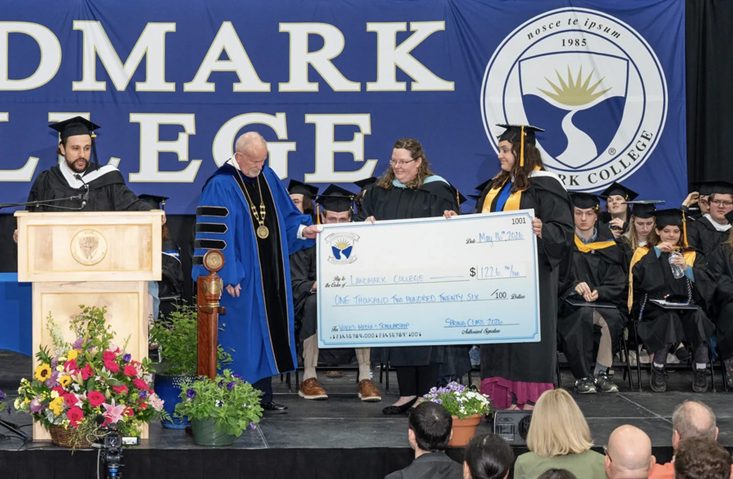 Two people present a large novelty to check to the president of the college, while a different person stands at a podium and speaks.