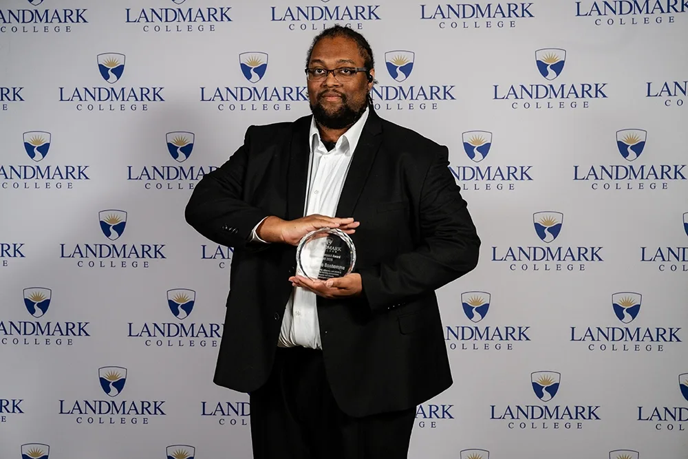 A man stands in front of a white background with the Landmark College logo while holding a glass award.