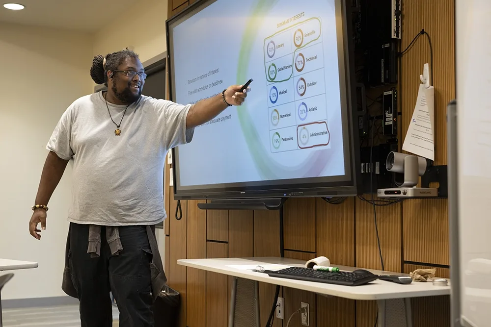 A man holds out a pointer while presenting research in front of a screen.