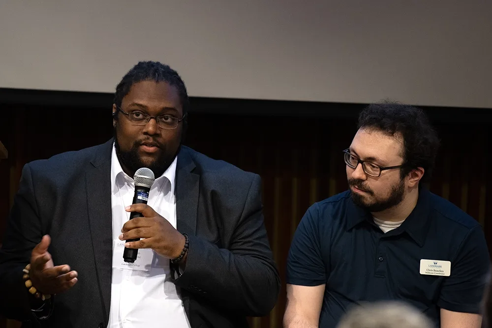 Two people, one holding a microphone, sit before a lecture hall.