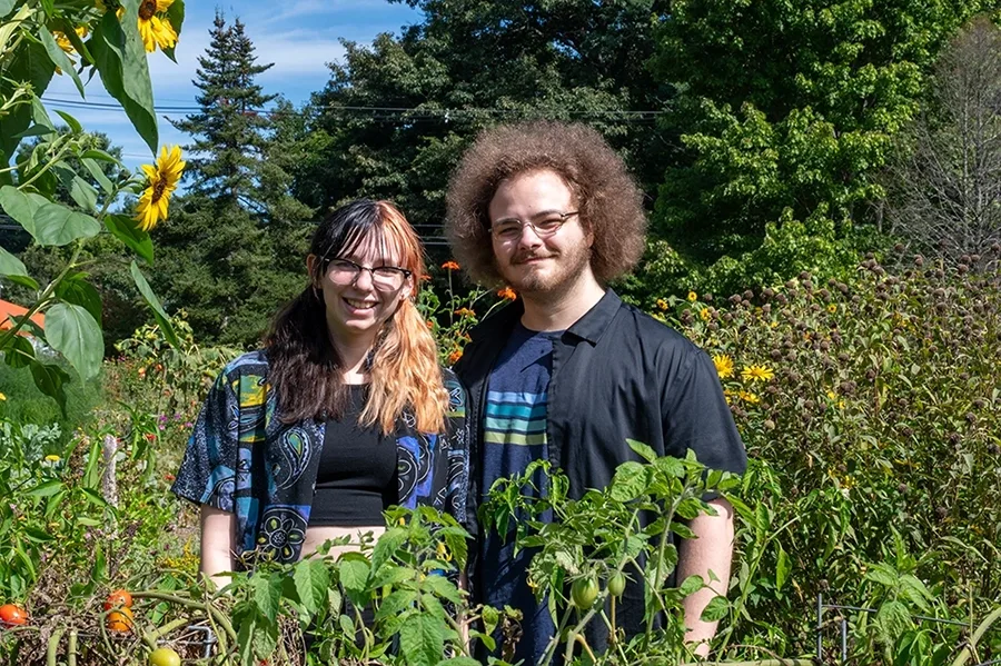 Two people stand in a field with flowers including sunflowers, and tomato plants, with trees in the background.