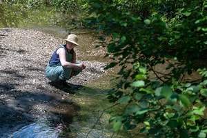 A person wearing a hat bends down near a body of water and some trees.