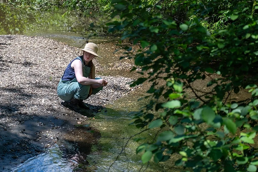 A person wearing a hat bends down near a body of water and some trees.