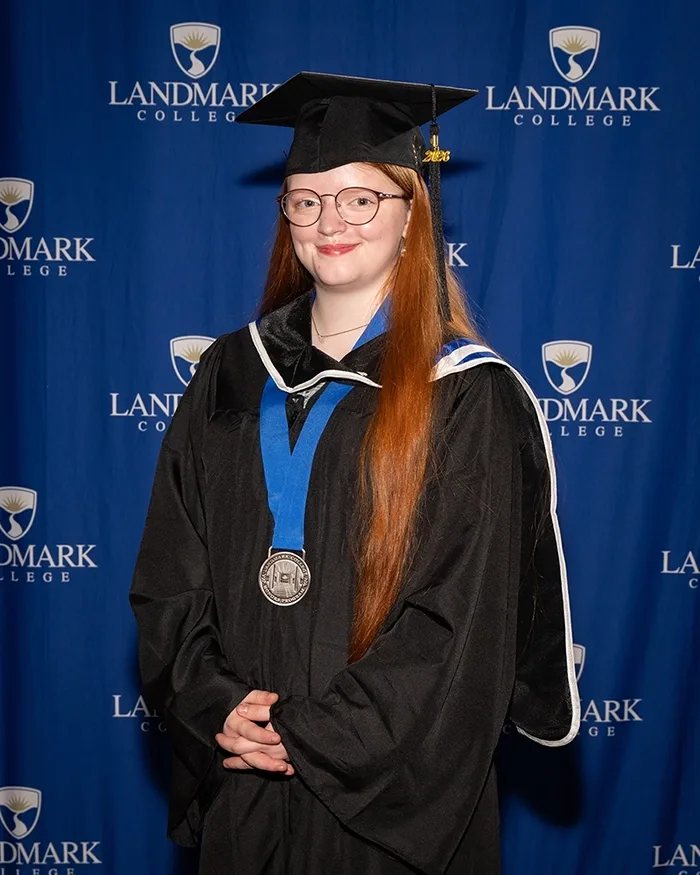 A person wearing a medal stands in a cap and gown in front of a blue background with the Landmark College logo on it.