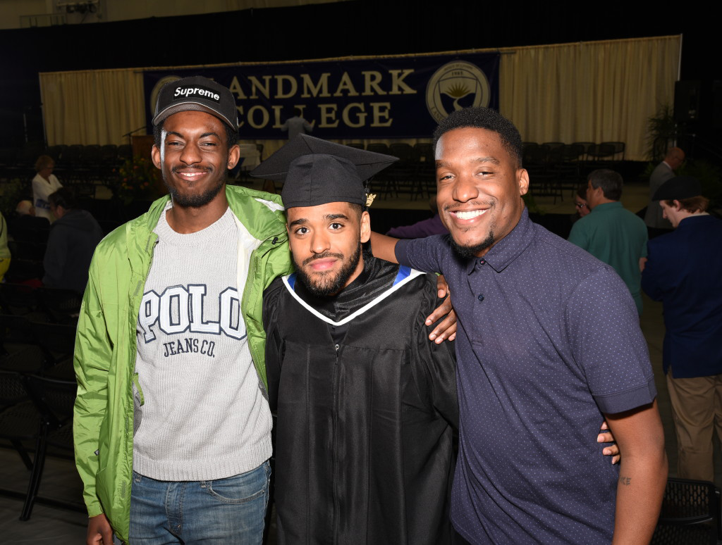Graduate in academic regalia poses with two friends in front of stage.