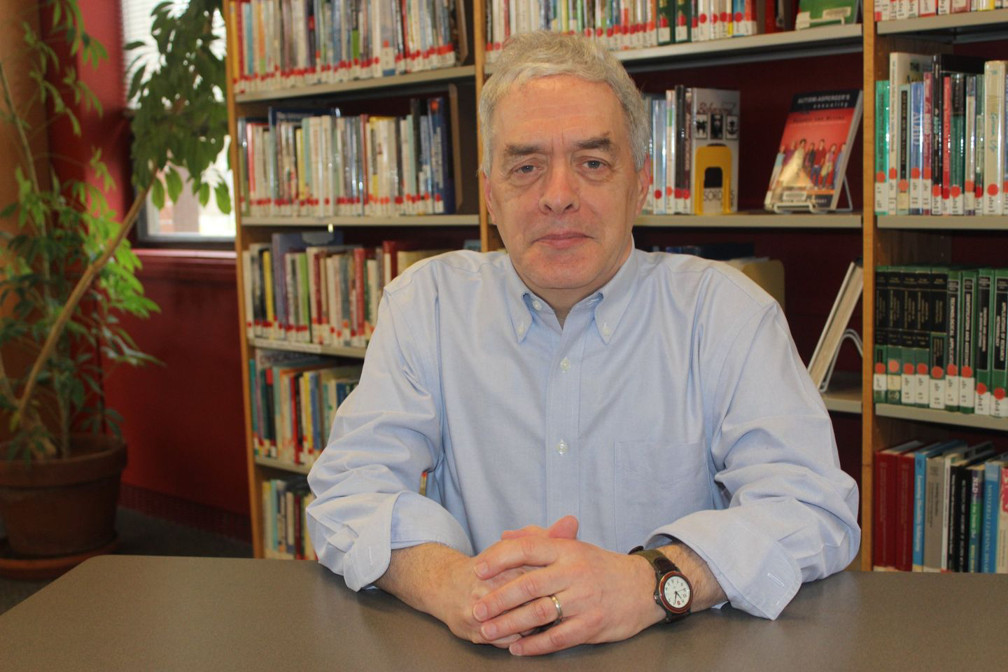 Gray-haired man in light blue button down shirt sits with hands folded at a table with library bookshelves behind him.