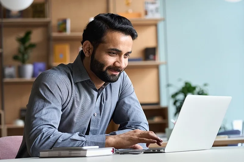 A man in a grey shirt working on a laptop at a desk with books and a smartphone.