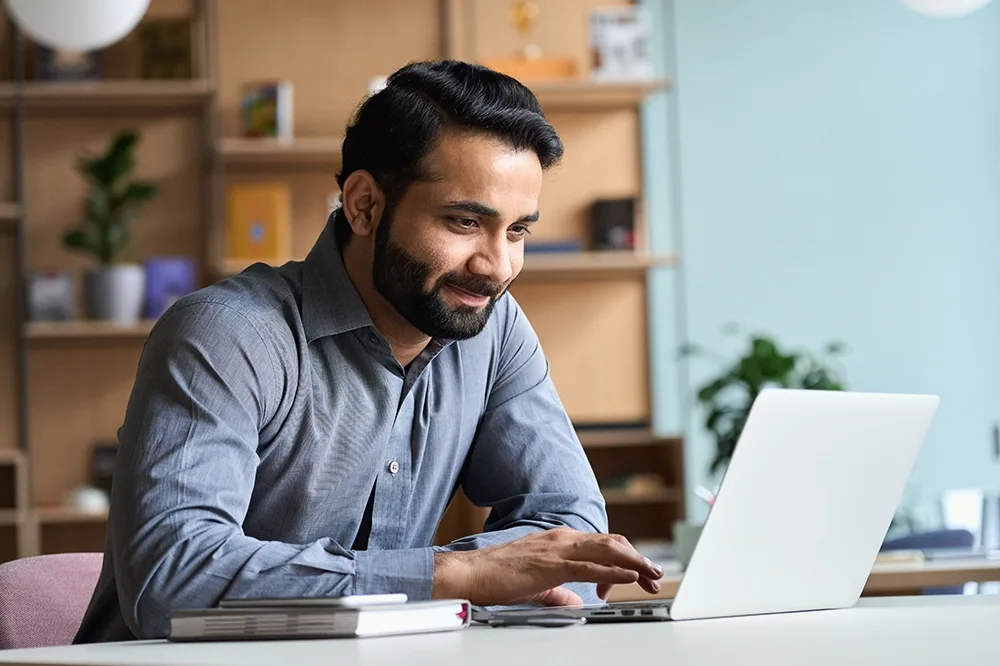 A man in a grey shirt working on a laptop at a desk with books and a smartphone.