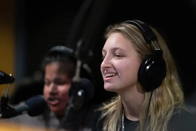 Two students on-air for a radio broadcast wearing headsets and talking into microphones.