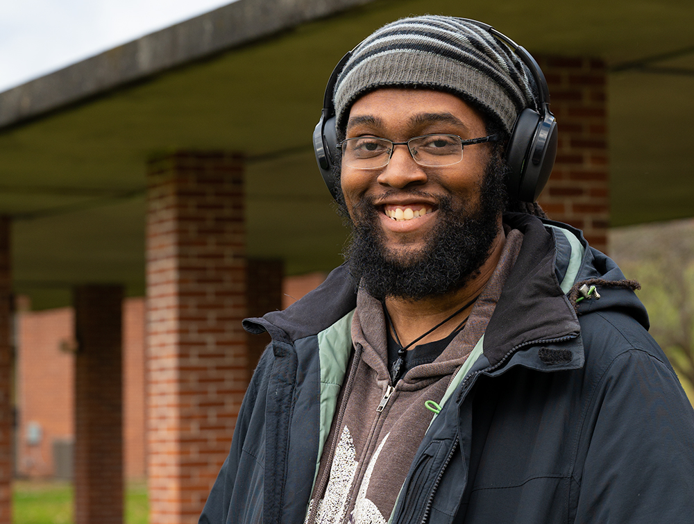 Photo of Anastasio Bonhomme smiling for camera on Landmark College quad. He is wearing a wool hat and headphones.
