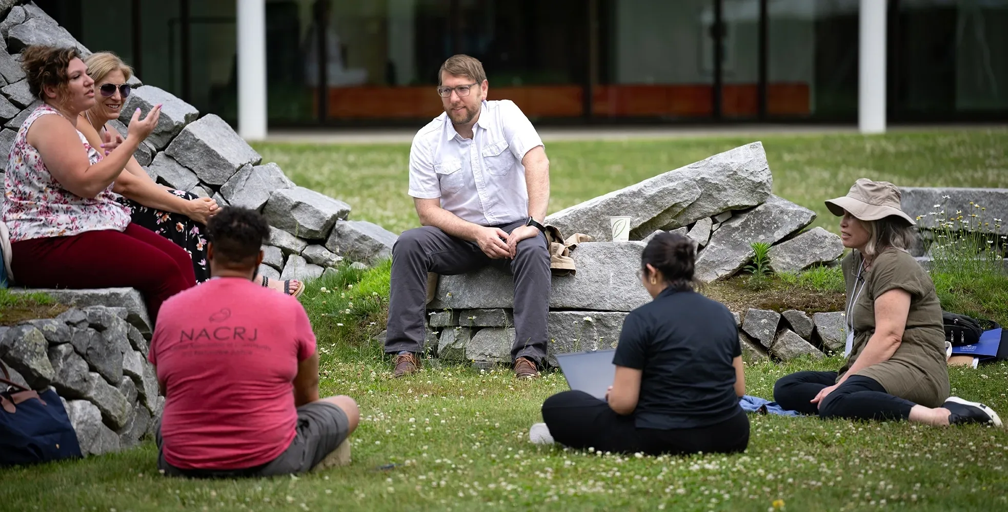 Six people sitting on the green lawn and shark stone sculpture outside of the STEM building for a summer institute workshop.