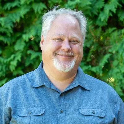 A headshot of Andrew Wilcox in front of arborvitae.